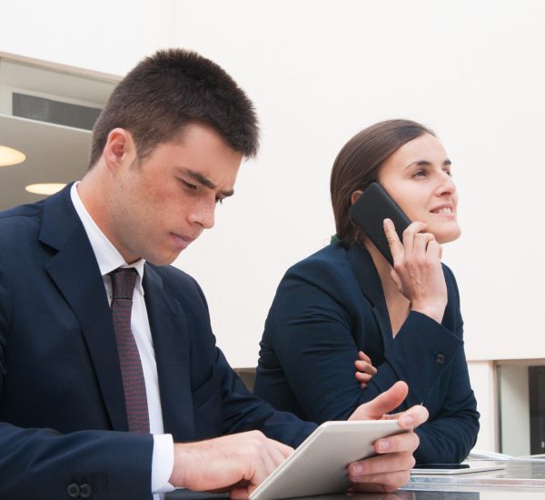 Colleagues using tablet and calling on phone at desk outdoors. Business man and woman wearing formal clothes and sitting at cafe table. Business and communication concept.
