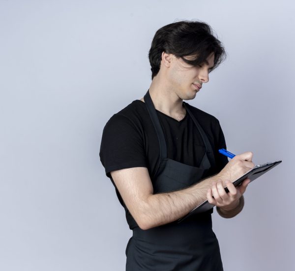 young handsome male barber in uniform writing something on clipboard isolated on white background
